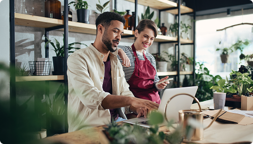 In a flower shop, a man gestures at a laptop as a woman observes, holding a phone in her hand. 