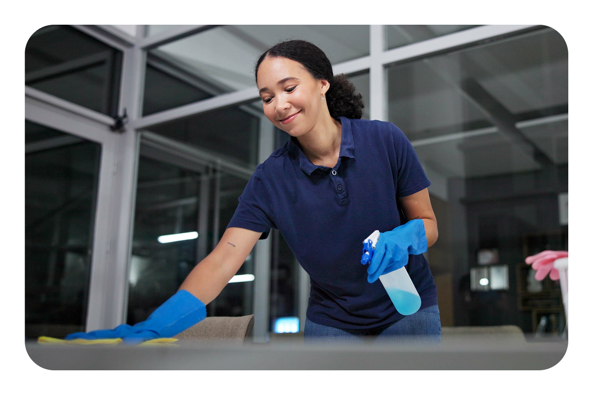Smiling woman in a blue polo shirt wearing light blue latex gloves cleaning a desk top 