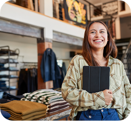 A woman inside a clothing store smiles while holding a mobile tablet device.