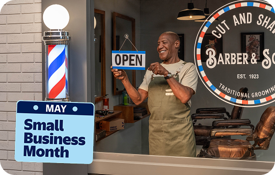 A man in a barber shop smiles as he flips his store sign to open. Text reads, May, Small Business Month.