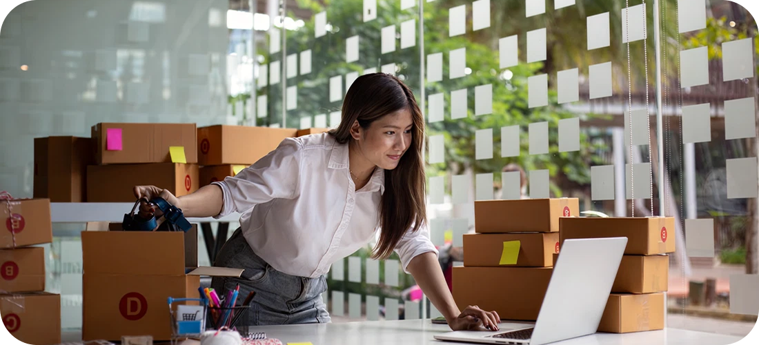 A woman multitasks, pulling inventory from a shipping box while working on a laptop.
