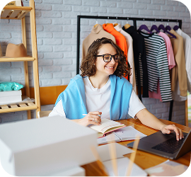A smiling woman with glasses working on a laptop at a desk with shipping boxes and a clothing rack in the background.