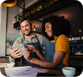 Two smiling business owners in striped aprons look at a digital tablet together in a modern café setting.